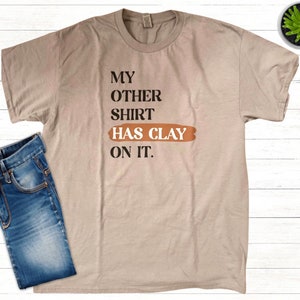 Puede incluir: Una camiseta beige con el texto "My other shirt has clay on it." impreso en negro. El texto "has clay" está impreso en un rectángulo marrón.