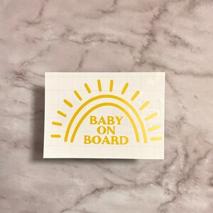 May include: Gold vinyl sticker with a sun design and the text "BABY ON BOARD".