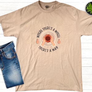 Puede incluir: Una camiseta beige con un gráfico de un torno de alfarería y el texto "Where there's a wheel there's a way".
