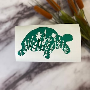 May include: A green vinyl decal of a turtle with white floral designs inside the silhouette. The turtle is walking to the right.