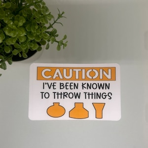 May include: A white sign with orange text that reads "CAUTION I'VE BEEN KNOWN TO THROW THINGS". Three orange silhouettes of objects are below the text: a round object, a vase-shaped object, and a funnel.