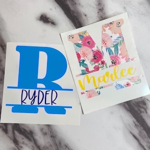 May include: Two personalized vinyl decals with floral designs. The first decal features a blue letter "R" with the name "Ryder" below it. The second decal features a floral letter "M" with the name "Marlee" below it.