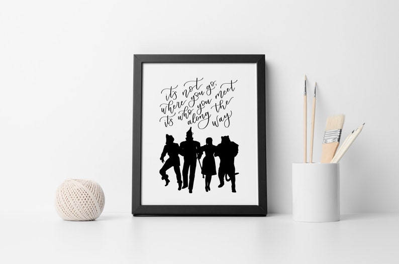 May include: Black and white printable wall art featuring a quote from the Wizard of Oz: "It's not where you go, it's who you meet along the way." The quote is written in a cursive font and is accompanied by a silhouette of the four main characters from the movie: Dorothy, the Scarecrow, the Tin Man, and the Cowardly Lion.