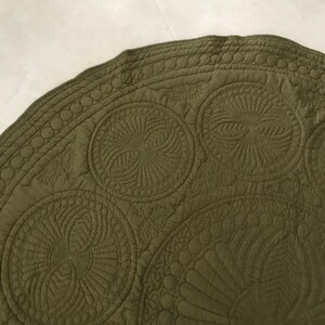 May include: Olive green circular quilted fabric with intricate stitched patterns. The design features large, spiraling circular motifs and detailed border stitching. The fabric has a matte finish and a soft texture.