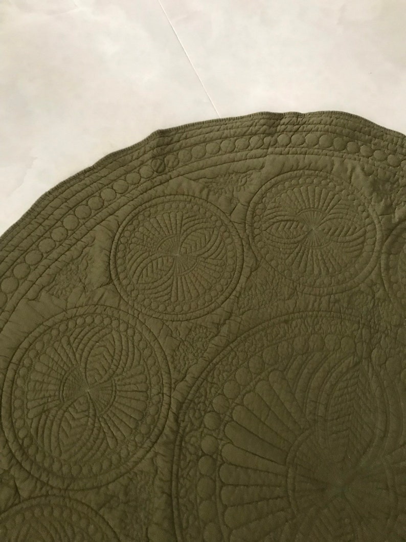 May include: Olive green circular quilted fabric with intricate stitched patterns. The design features large, spiraling circular motifs and detailed border stitching. The fabric has a matte finish and a soft texture.