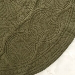May include: Close-up of a section of an olive green quilted textile. The fabric features intricate circular patterns with detailed stitching, including spirals and concentric circles. The edge has a border of small circles.