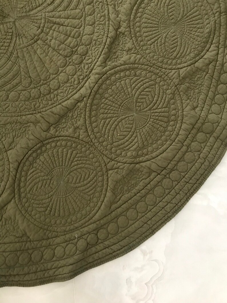 May include: Close-up of a section of an olive green quilted textile. The fabric features intricate circular patterns with detailed stitching, including spirals and concentric circles. The edge has a border of small circles.