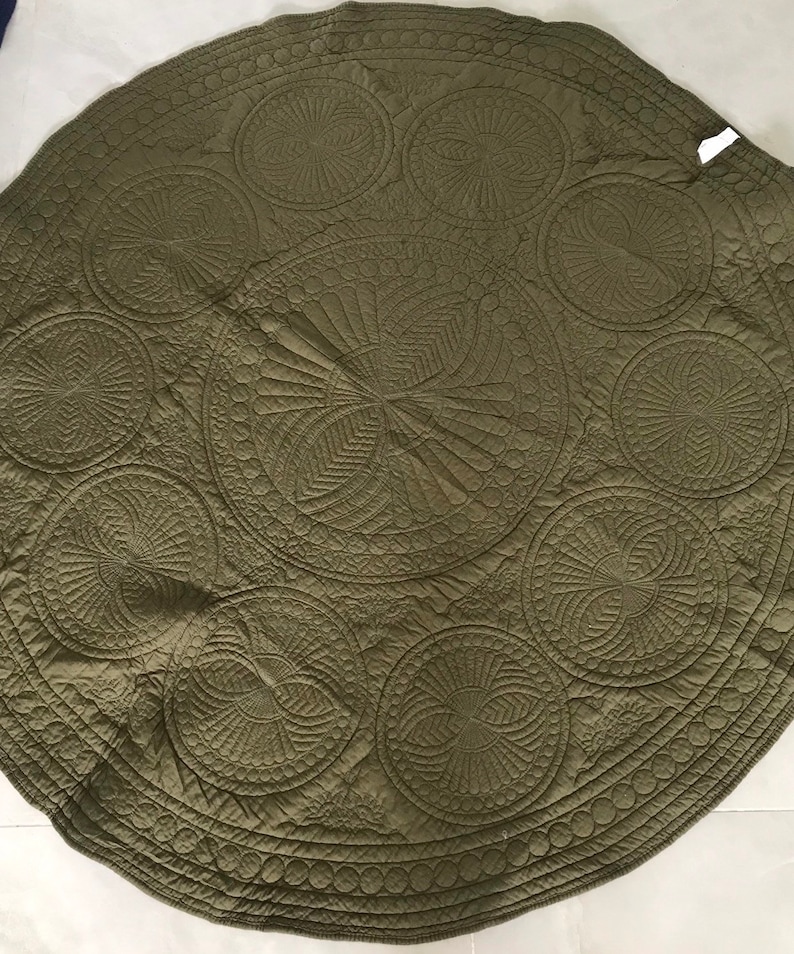 May include: Olive green, round quilted textile with intricate stitched circular patterns. The design features various sizes of circles with detailed internal designs, creating a textured surface. The edges are scalloped.