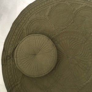 May include: Olive green round quilted mat and matching round pillow. The mat features a detailed circular pattern with a scalloped edge. The pillow has a similar design. The set is made of a soft, textured fabric.