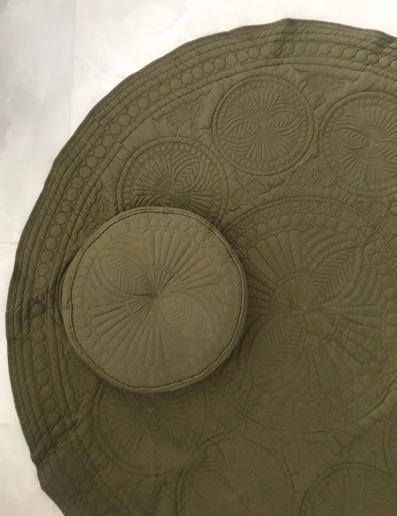 May include: Olive green round quilted mat and matching round pillow. The mat features a detailed circular pattern with a scalloped edge. The pillow has a similar design. The set is made of a soft, textured fabric.