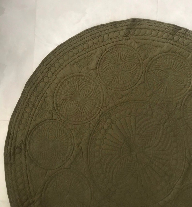 May include: Olive green, circular quilted fabric with intricate stitched circular and fan-like patterns. The edge features a repeating circle design. The fabric has a soft, matte finish and appears to be made of cotton or a similar material.