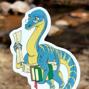 Może przedstawiać: Niebieski i żółty dinozaur w okularach trzyma zwój i książkę. Dinozaur ma zieloną książkę pod pachą i czerwoną wstążkę zwisającą z książki.