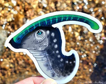 May include: A die-cut sticker featuring a Parasaurolophus dinosaur head in profile. The dinosaur has a grey, speckled neck and head with a bright blue eye. A long, curved, green and blue crest extends from the back of the head. The sticker has a white border.