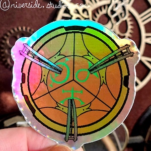 Peut inclure: Un autocollant holographique avec un motif circulaire dans les tons vert, or et orange. Le motif comprend un symbole central à trois points et deux épées croisées.