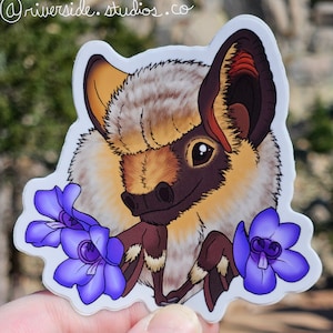 May include: A sticker featuring a cartoon illustration of a brown and white bat with large ears and a fluffy appearance. The bat is surrounded by three purple flowers with yellow centers.
