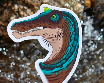 May include: A sticker of a blue and brown dinosaur with a green crest. The dinosaur has a toothy grin and is set against a blurry background of water.