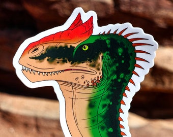 May include: A sticker featuring a close-up illustration of a dinosaur head. The dinosaur has a red crest, green scales, and a white background. The sticker is printed on white paper.