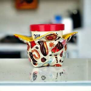 Op de afbeelding: Een kleurrijke stoffen ijsbekerhouder met een rood, wit en geel ontwerp met verschillende gebakjes en desserts. De houder houdt een witte ijscontainer met een rood deksel.