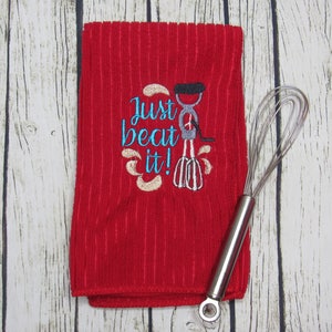 Puede incluir: Toalla de cocina roja con el texto "Just beat it!" bordado en azul turquesa con una imagen estilizada de una batidora de mano en gris y negro.