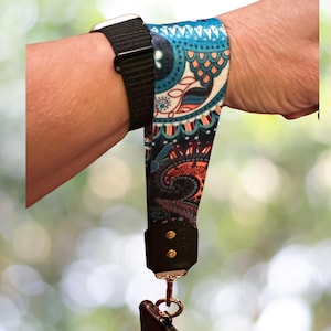 Könnte beinhalten: Ein gemustertes Handgelenkband mit schwarzer Lederbasis und goldfarbenen Nieten. Das Band weist ein farbenfrohes Paisley-Design in Blau-, Orange- und Schwarztönen auf. Ein schwarzes Uhrenarmband ist ebenfalls sichtbar.