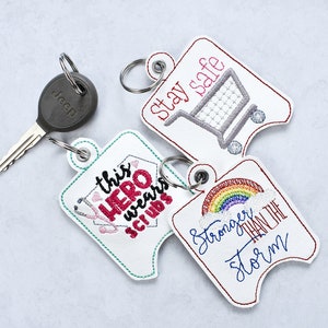 May include: Three white keychains with colorful embroidery. One keychain says "This Hero Wears Scrubs" with a stethoscope and a heart. Another keychain says "Stay Safe" with a shopping cart. The third keychain says "Stronger Than The Storm" with a rainbow and a cloud.
