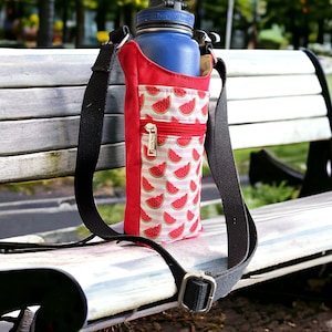 May include: A red and white watermelon print water bottle carrier with a black adjustable strap. The carrier is sitting on a wooden bench.
