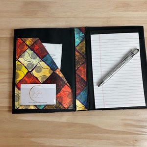 May include: A black leather portfolio with a colorful patterned fabric insert. The portfolio has a pocket for a business card and a notepad with lined paper. A silver pen with the text "The Creative Lady Co." is on the notepad.