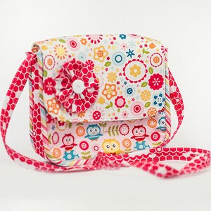 Pu&ograve; includere: Una piccola borsa di tessuto rosa e bianco con una stampa floreale e un accento di fiore rosso. La borsa ha una lunga tracolla rosa con un motivo a nido d'ape.