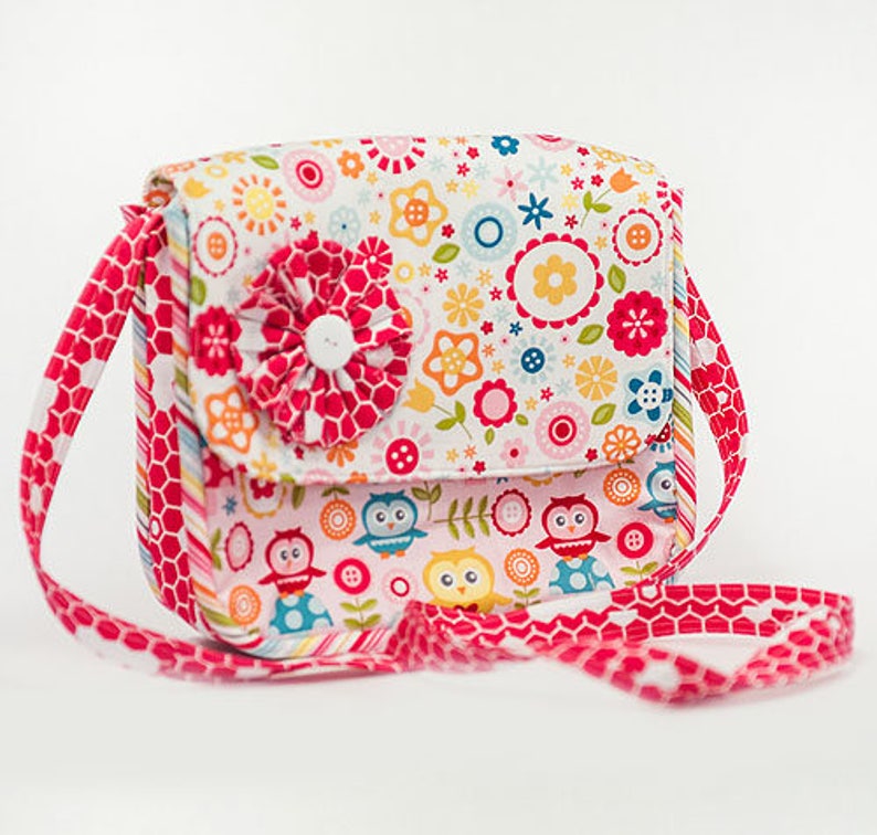 Pu&ograve; includere: Una piccola borsa di tessuto rosa e bianco con una stampa floreale e un accento di fiore rosso. La borsa ha una lunga tracolla rosa con un motivo a nido d'ape.