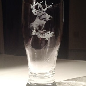 May include: A clear glass beer pilsner with etched images of a deer and a fish. The deer has large antlers and the fish is a bass. The glass is tapered and has a wide base.