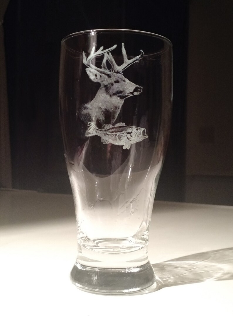May include: A clear glass beer pilsner with etched images of a deer and a fish. The deer has large antlers and the fish is a bass. The glass is tapered and has a wide base.