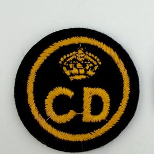 May include: A circular black patch with gold embroidery. It features a gold crown above the gold letters "CD". A gold border surrounds the letters and crown, creating a detailed design.