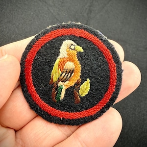 May include: A circular embroidered patch featuring a black background with a red border. The centre displays a detailed bird with white, orange and yellow plumage perched on a brown branch with a single green leaf, rendered in thread.