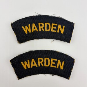 May include: Two black fabric patches, each with the word "WARDEN" in yellow lettering. The patches are curved and have a distressed edge. They are likely uniform insignia.
