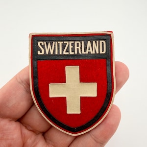 以下が含まれることがあります： スイス国旗のデザインが施されたシールド型のパッチ。赤い背景に白い十字、黒い縁取り、そして白い文字で「SWITZERLAND」と書かれています。