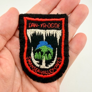 May include: A black embroidered patch with red trim and white text reading "DAN-YR-OGOF" and "SWANSEA VALLEY CAVES". The patch features a blue and green mountain scene with white teeth-like shapes.