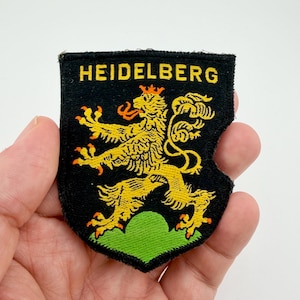 May include: Embroidered patch with the word "HEIDELBERG" in yellow on a black background. It features a yellow lion rampant with a red crown and accents, above a green base.