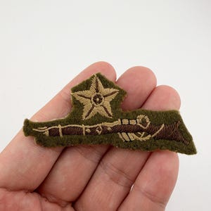 May include: An embroidered military badge in olive green, brown, and tan. The badge features a star and rifle design. The patch is rectangular with a star at the top and a rifle design below. The badge is held in a hand.