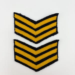 May include: Two yellow and black chevron patches. Each patch features three yellow stripes on a black background, arranged in a V-shape. These patches are likely for military or uniform use.
