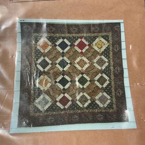 May include: A square quilt pattern featuring a grid of diamond-shaped blocks in various colors including red, yellow, green, and blue, set against a brown background with a dark brown floral border, all enclosed within a clear plastic packaging sleeve.