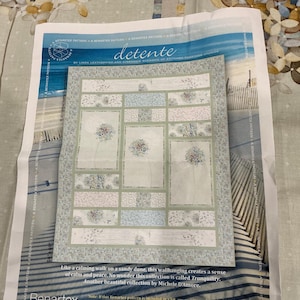 May include: A quilt pattern titled "detente" by Benartex, featuring a floral design in soft blues and greens. The pattern is displayed on a printed sheet with a beach scene background. The text on the sheet mentions "Tranquility".