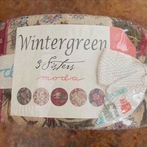 May include: A fabric roll wrapped in clear plastic, featuring a variety of floral and patterned fabrics. The label reads "Wintergreen 3 Sisters moda" with a decorative bow. The fabric colors include red, green, and beige.