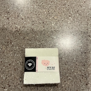 May include: A stack of light green fabric squares with white polka dots, wrapped with a label. The label features the text "SUGAR by Sarah Watts" and a pink cloud illustration. The fabric measures 2.5 inches.