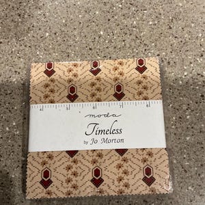 May include: A stack of square fabric swatches featuring a tan background with a repeating pattern of small brown flowers and dark red geometric shapes. A white paper band labeled "moda Timeless by Jo Morton" with a 5-inch ruler is wrapped around it.
