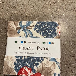 May include: A stack of square fabric swatches featuring a floral pattern with blue, red, and beige tones. A white paper band wraps around the center, printed with the text "moda", "GRANT PARK", and "by Minick & Simpson for moda".