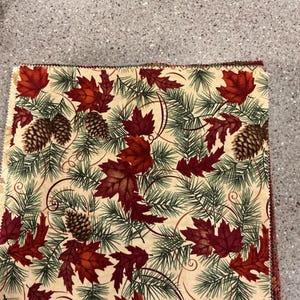 May include: A stack of fabric squares with a fall-themed pattern. The design features red maple leaves, green pine needles, and brown pine cones on a cream-colored background. The fabric is likely cotton or a similar material.
