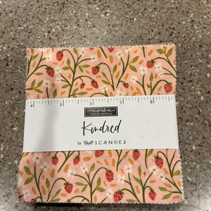 May include: A stack of fabric squares with a strawberry and floral pattern. The fabric is pink with red strawberries, white flowers, and green leaves. The label reads "Kindred" by 1Canoe2.