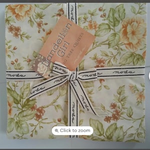 May include: A square fabric bundle featuring a floral pattern in cream, peach, and green. The bundle is tied with a cream ribbon printed with "moda" and a bow. A tag reads "Dandelion Girl" and "Fig Tree Quilts."