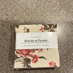 May include: A stack of fabric squares with a floral pattern in cream, red, and brown. The fabric is labeled "ATELIER DE FRANCE" by French General for moda. A ruler is placed on top of the fabric.