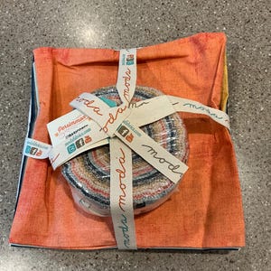 May include: A fabric bundle wrapped in orange fabric and tied with a ribbon that says "moda". The bundle contains a roll of fabric strips and is labeled "Persimmon Basics". The fabric has a variety of colors and patterns.